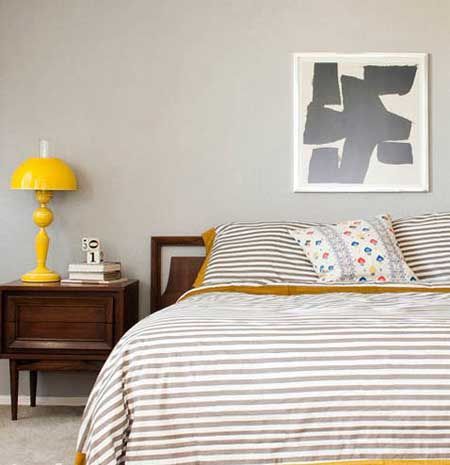 卧室墙面大面积涂刷了浅灰色的墙漆，所有的软装饰全部都同色系，暖色调的黄色边围围绕着条纹床品与同样明媚无比的黄色床头灯，组合在一起营造出卧室温和亲近的性格